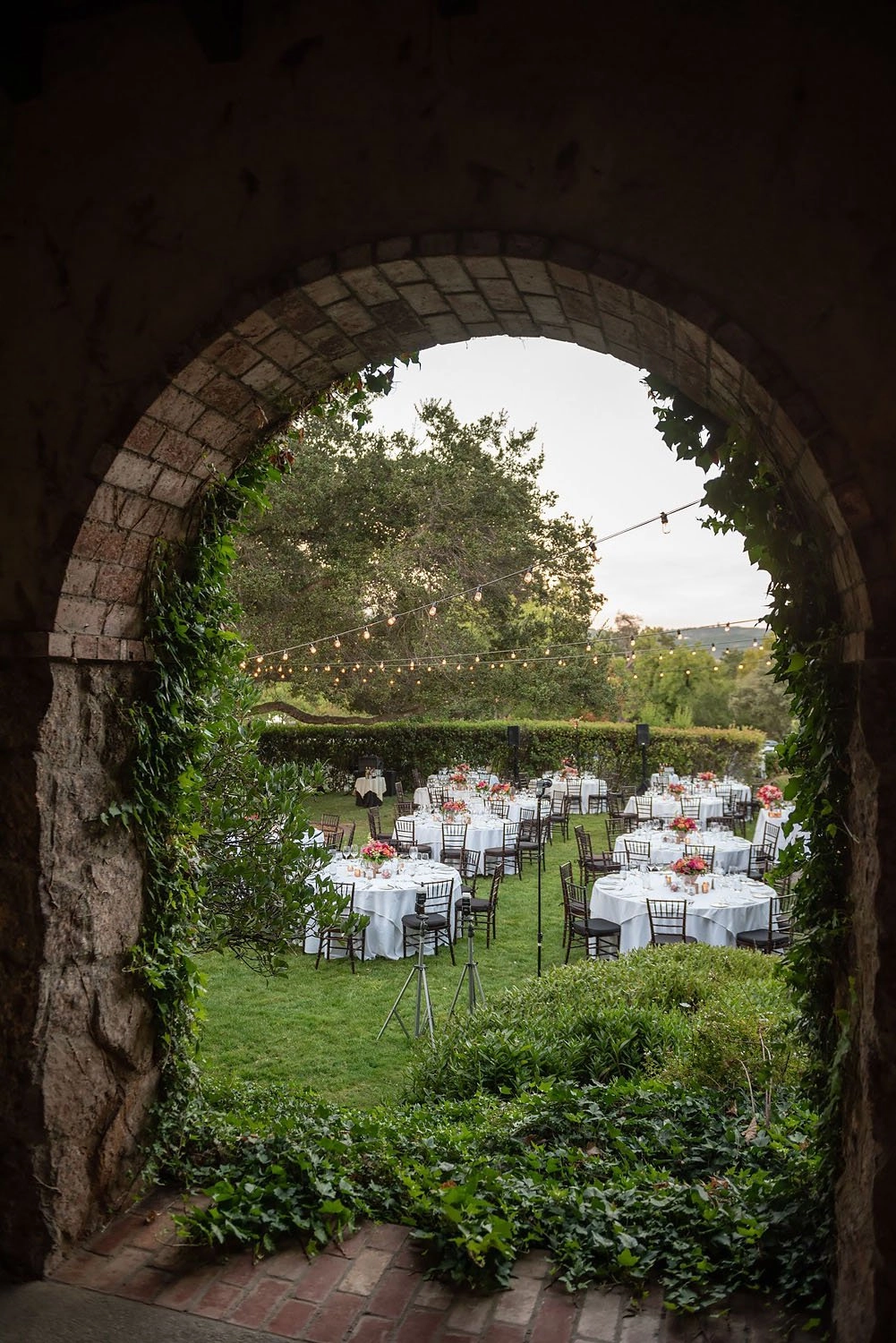 Garden reception seen through a stone archway with bistro lights and coral centerpieces